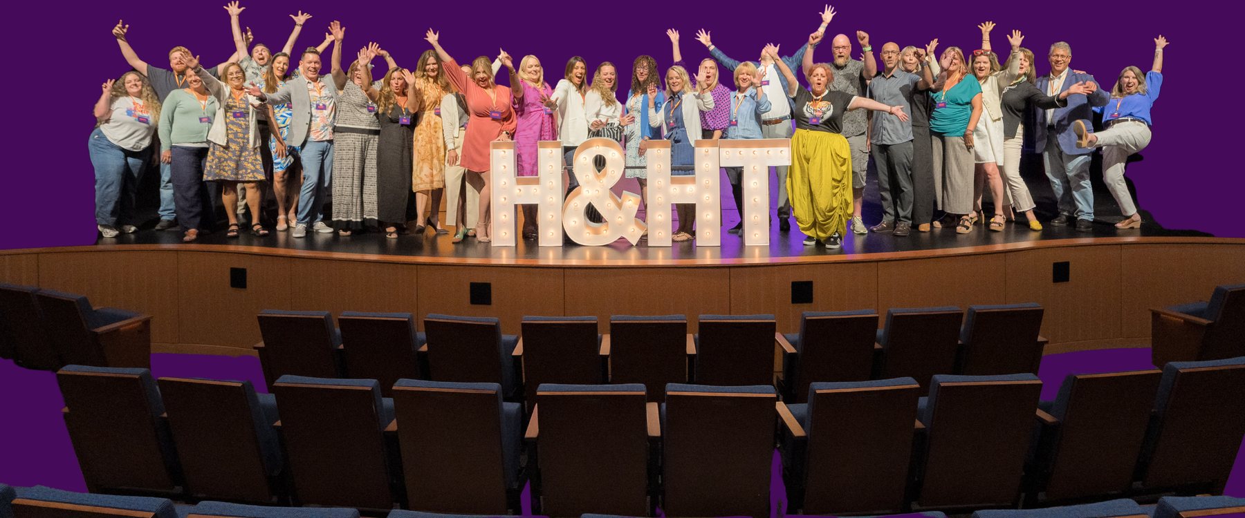 Participants of the inaugural Hosts & Home Teams Field Lab on stage in Portland, arms raised behind illuminated H&HT marquee letters